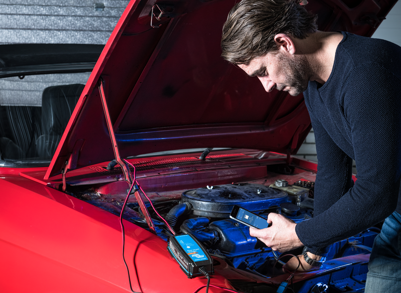 Man working on a red car with the hood open, using a device to check the battery.